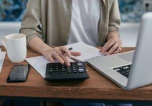 Person Writing on Notepad Using a Calculator and Laptop Sitting at a Wooden Desk